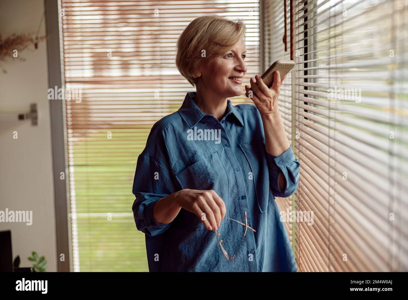 Donna di mezza età che registra un messaggio audio o utilizza la funzione Voicemail mobile, in piedi vicino alla finestra Foto Stock