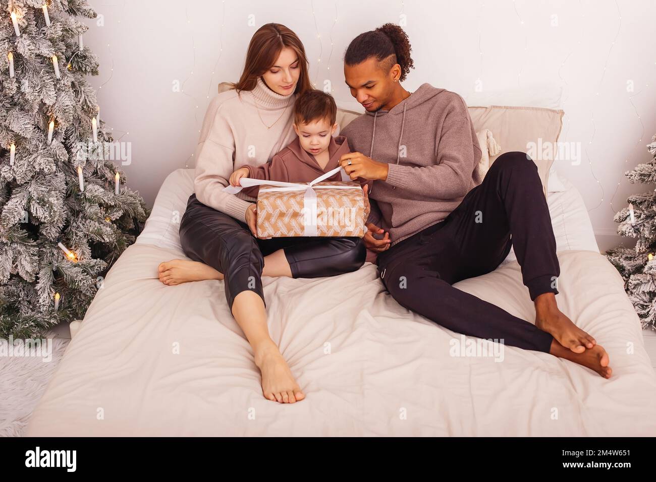 Una famiglia stilysh sta sedendosi su un letto, in un interno di Natale, slegando un nastro su una scatola marrone del regalo Foto Stock