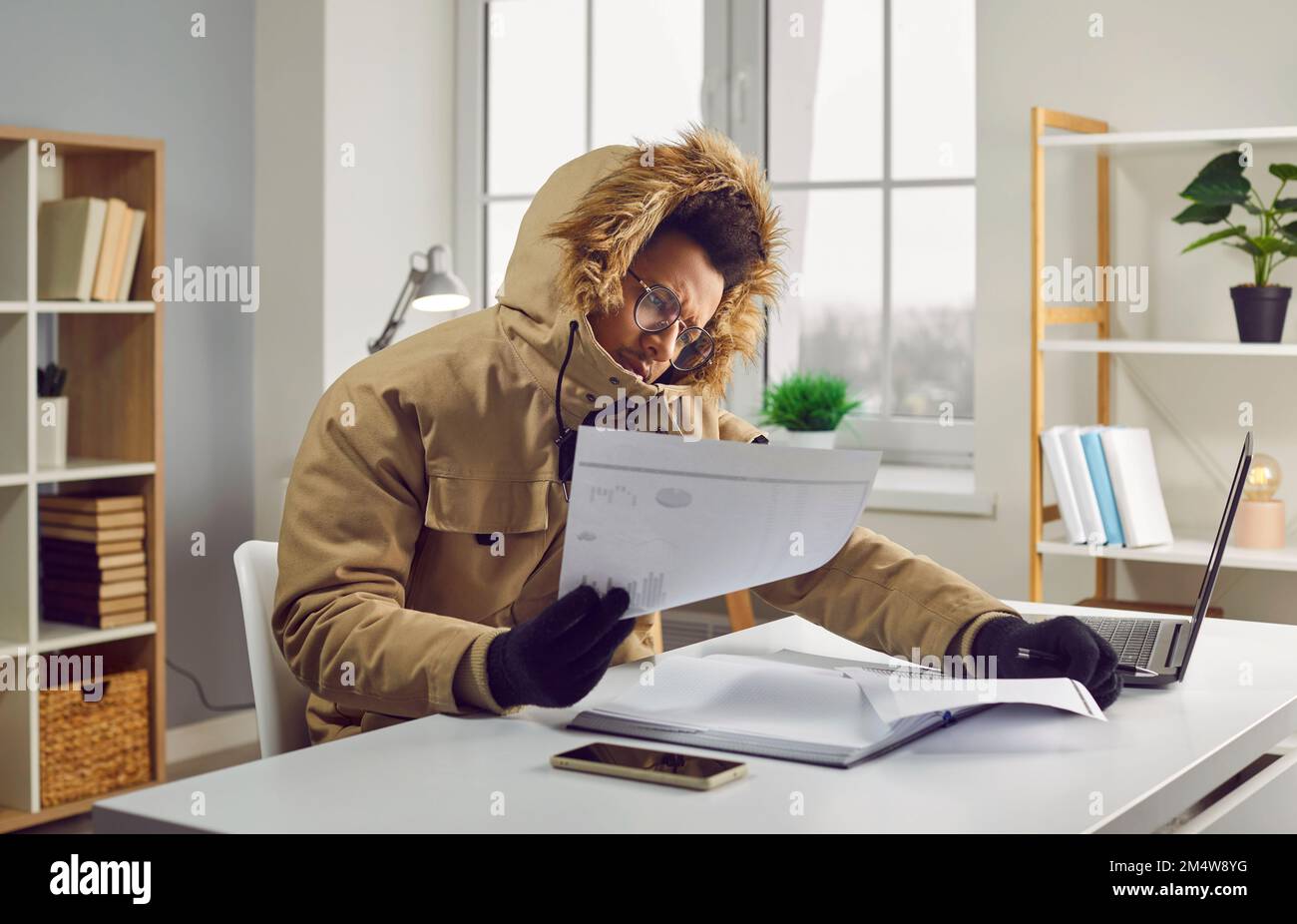Uomo in un abbigliamento invernale guardando bollette di riscaldamento mentre seduto alla sua scrivania nella sua casa fredda Foto Stock