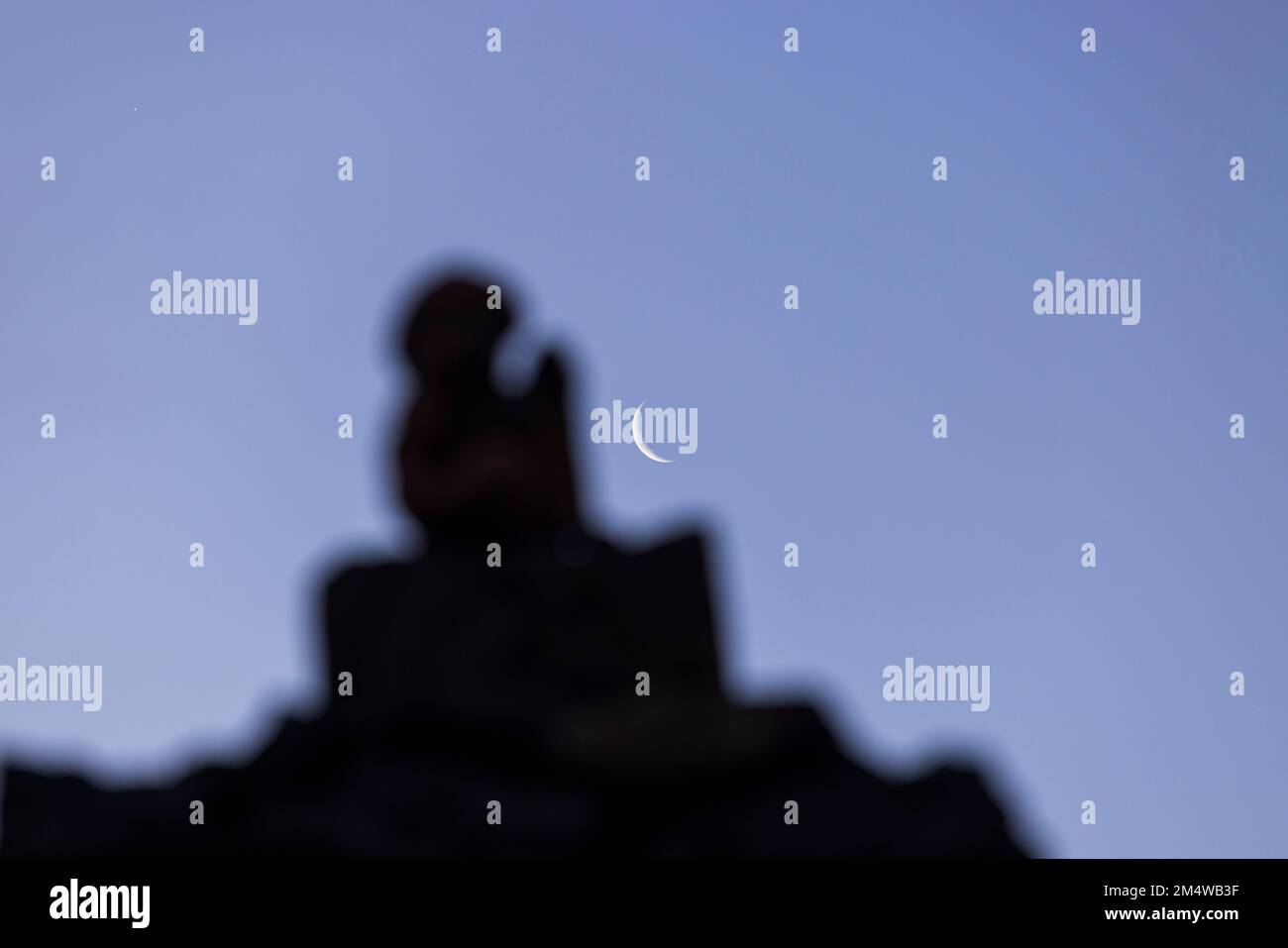 Luna di Mezzaluna in un cielo blu con la silhouette scura di una statuetta fuori fuoco in primo piano, Caserio Los Partidos, El Tanque, Tenerife, CA Foto Stock