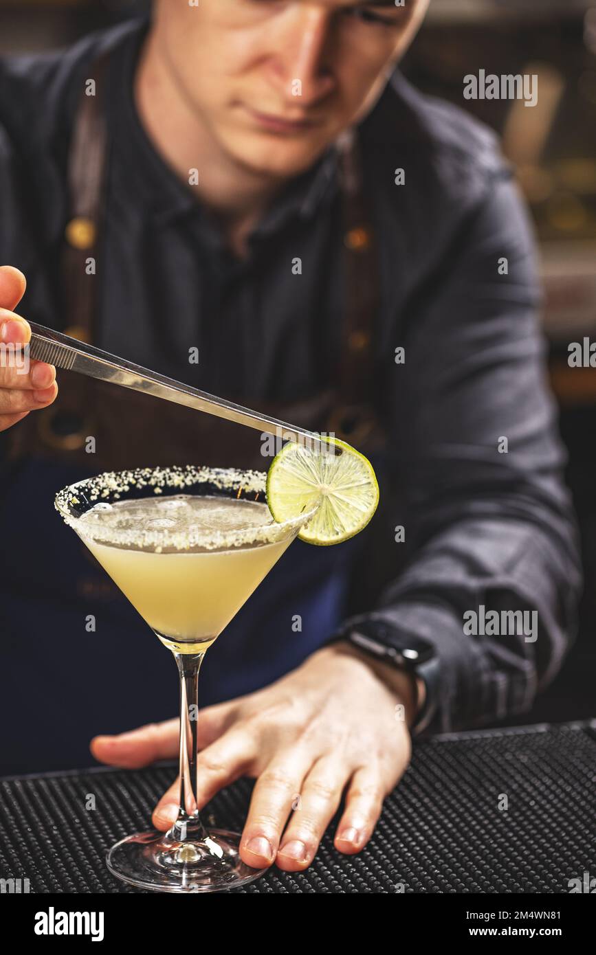 Bicchiere Martini con cocktail luminoso sul banco del bar e mano del barman tiene una fetta di limone con pinzette e decora il bicchiere con esso Foto Stock
