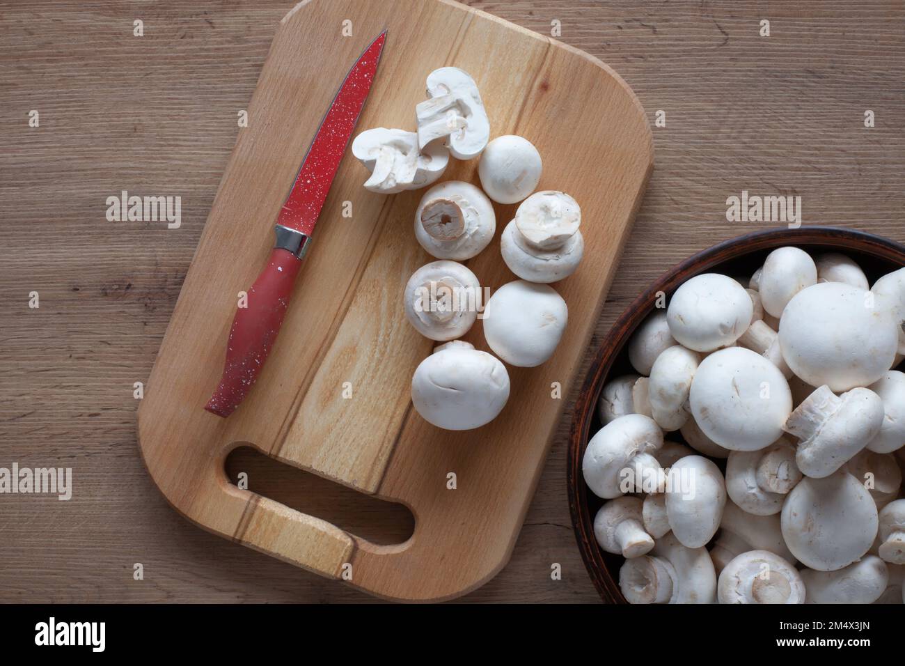 Champignons freschi sul tavolo, in una ciotola e su un asse di legno con un coltello rosso, vista dall'alto degli ingredienti vegetali per la cottura. Foto Stock