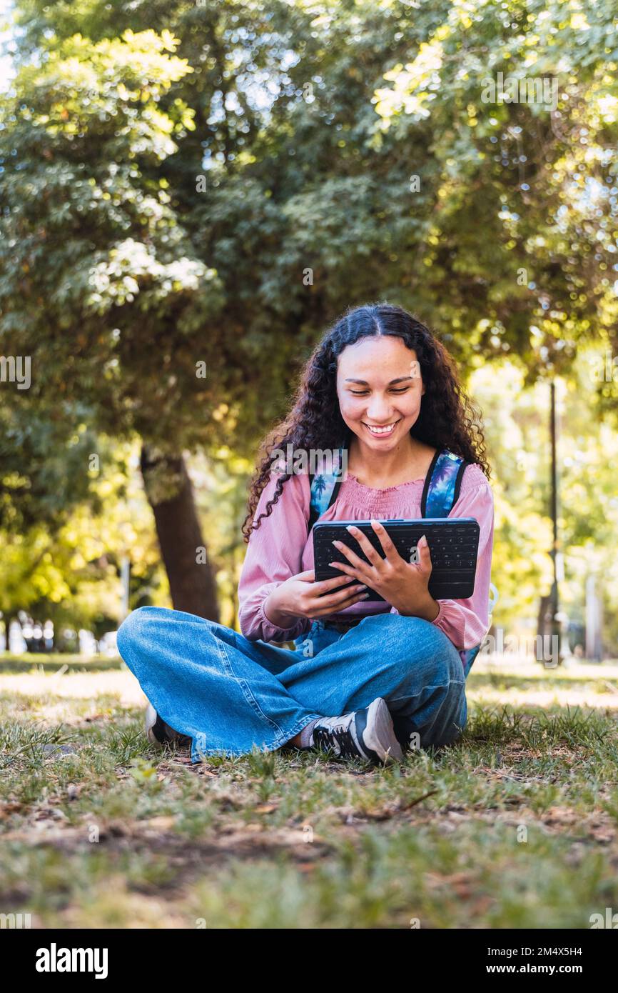 Studentessa universitaria nera che sorride e usa un tablet seduto all'aperto in un parco sull'erba Foto Stock