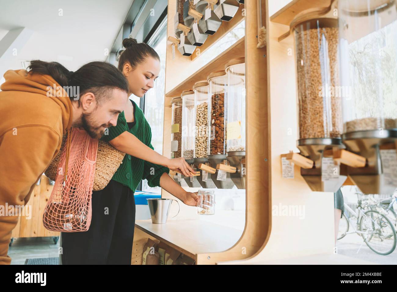 Uomo che guarda la donna che riempie il vasetto di mason sotto il dispenser di cibo a zero rifiuti negozio Foto Stock