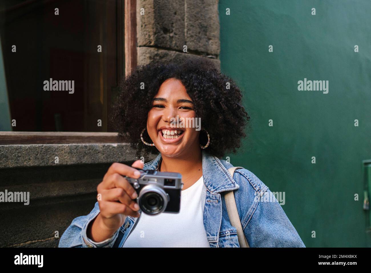 Giovane donna allegra con capelli ricci che tiene la macchina fotografica analogica davanti al muro Foto Stock