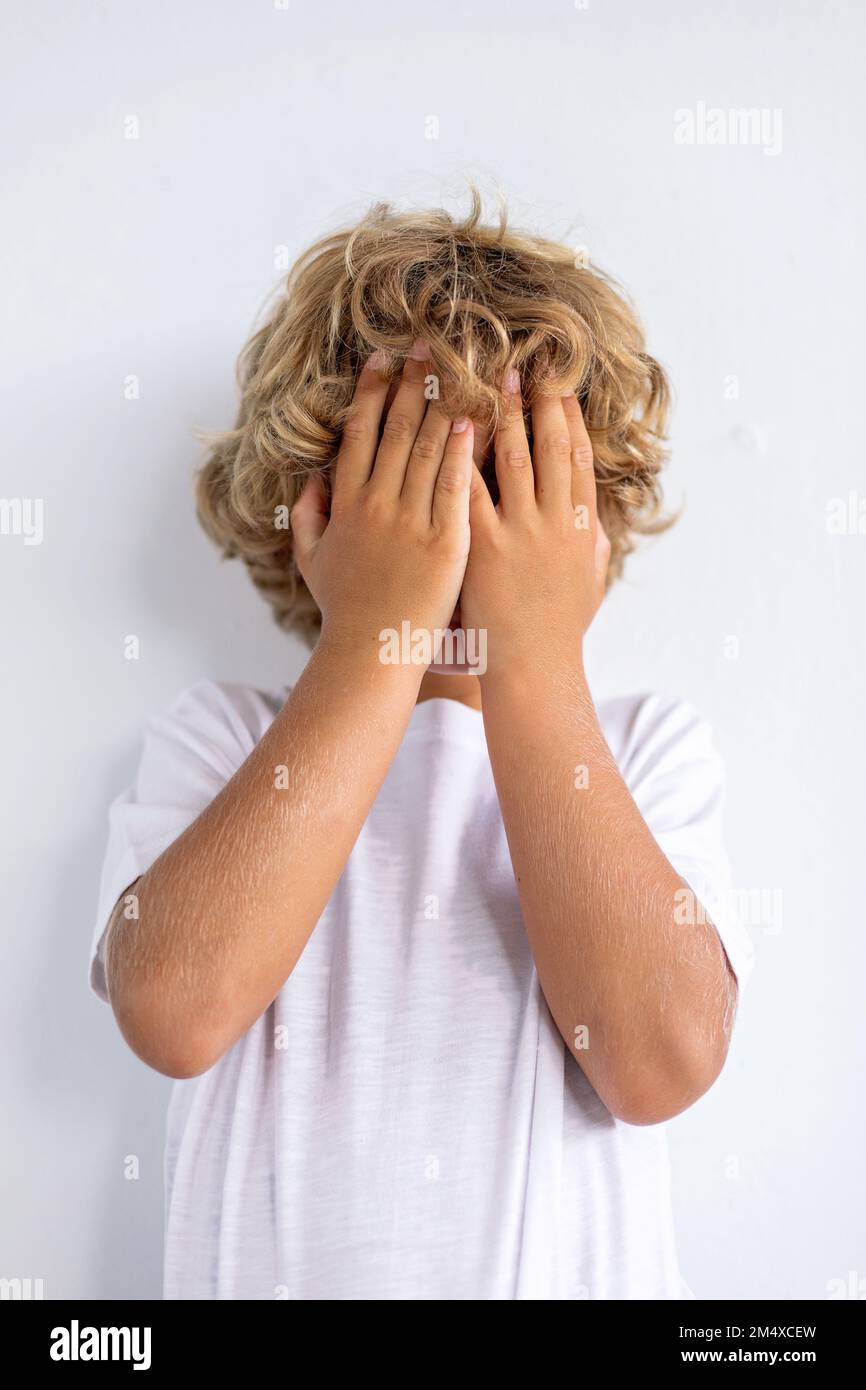 Ragazzo timido che copre il viso con le mani su sfondo bianco Foto Stock