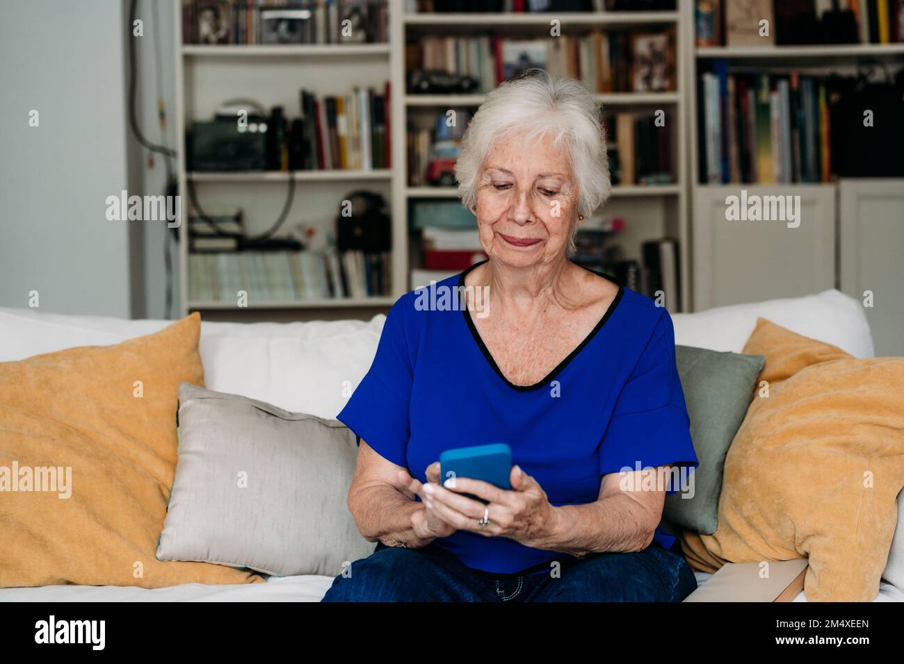 Donna anziana sorridente che usa lo smartphone sul divano del soggiorno Foto Stock