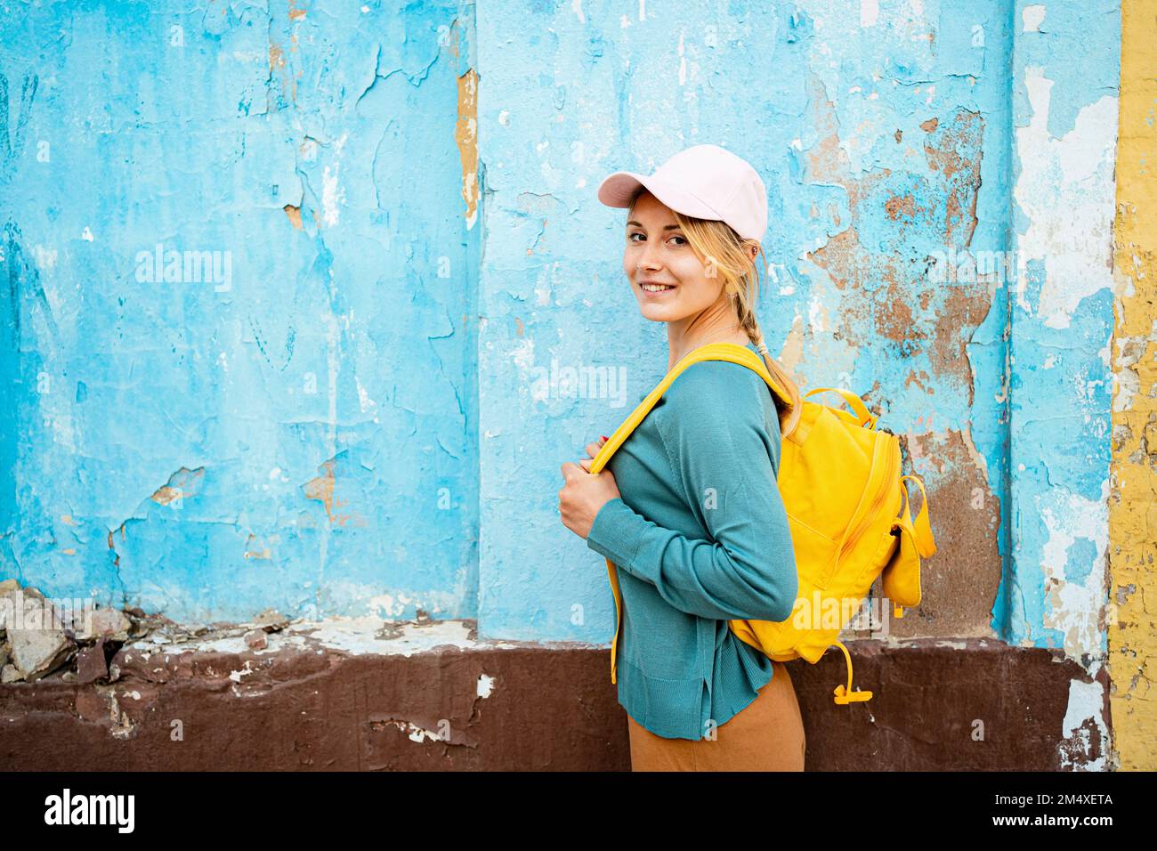 Donna sorridente con zaino giallo in piedi da una parete incrinata Foto Stock
