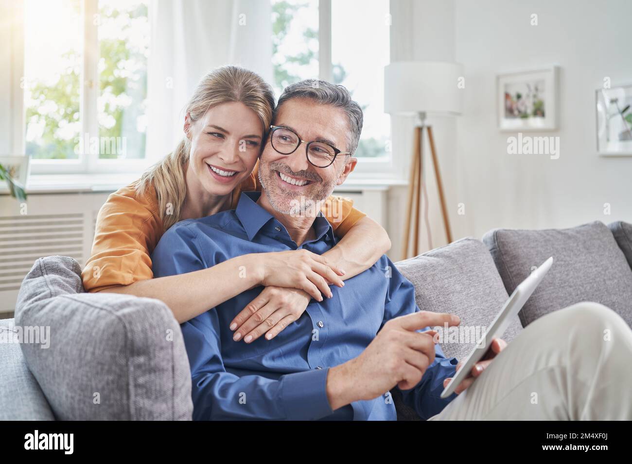 Donna felice che abbraccia un uomo che tiene il tablet PC da dietro sul divano di casa Foto Stock