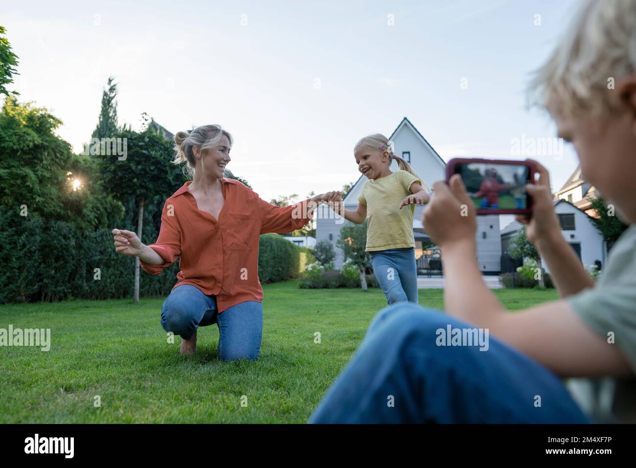 Ragazzo fotografando la madre ballando con la ragazza attraverso il telefono cellulare in giardino Foto Stock