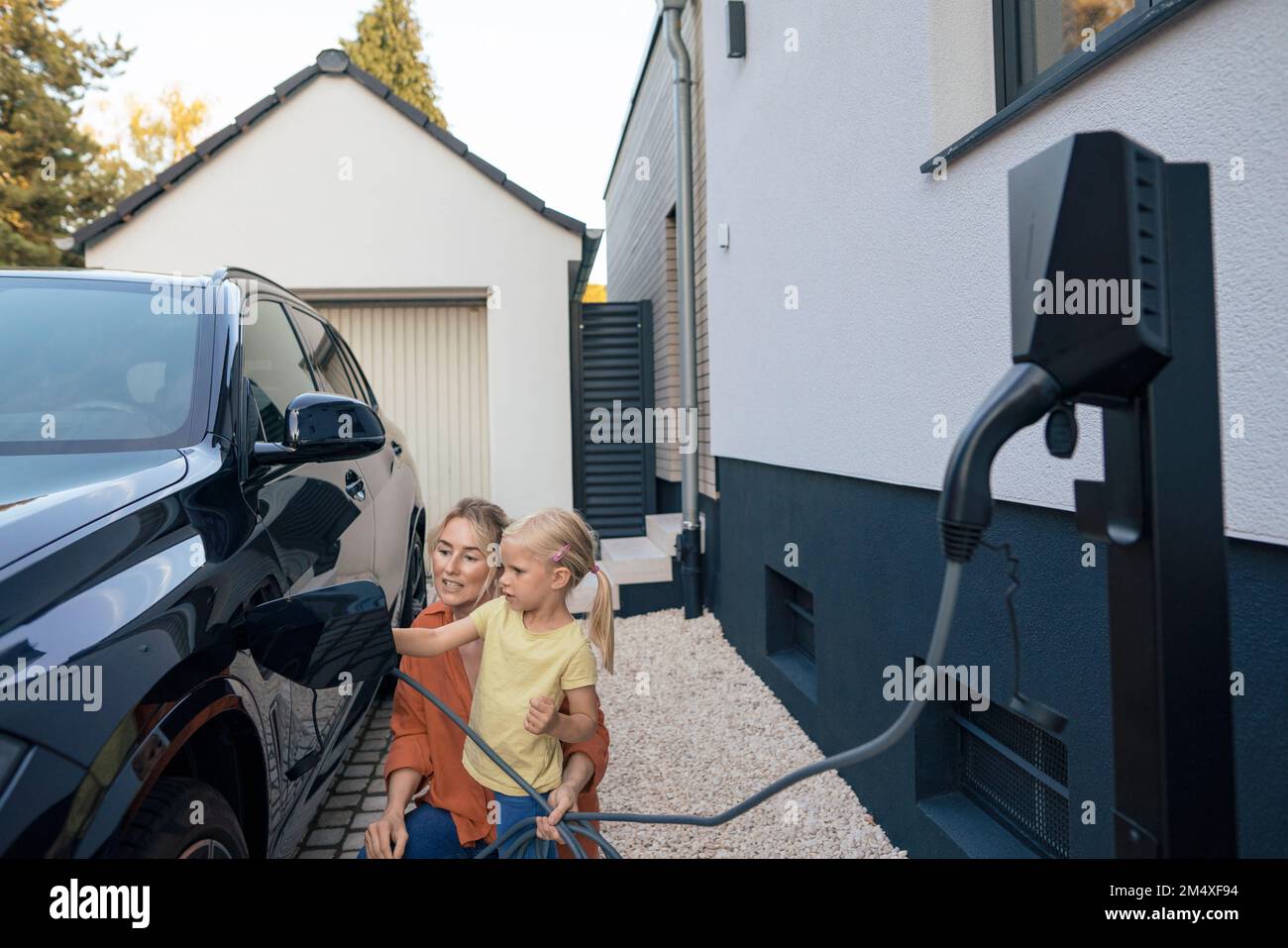Madre che guida la figlia per caricare l'auto elettrica nel cortile anteriore Foto Stock