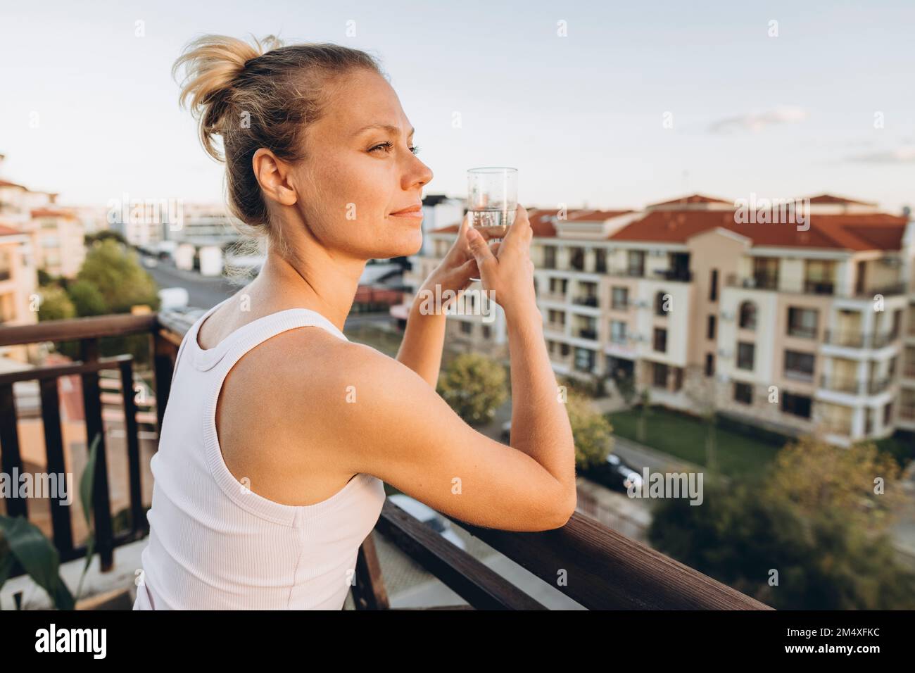 Donna appoggiata su ringhiera di terrazza sul tetto che tiene vetro di acqua Foto Stock