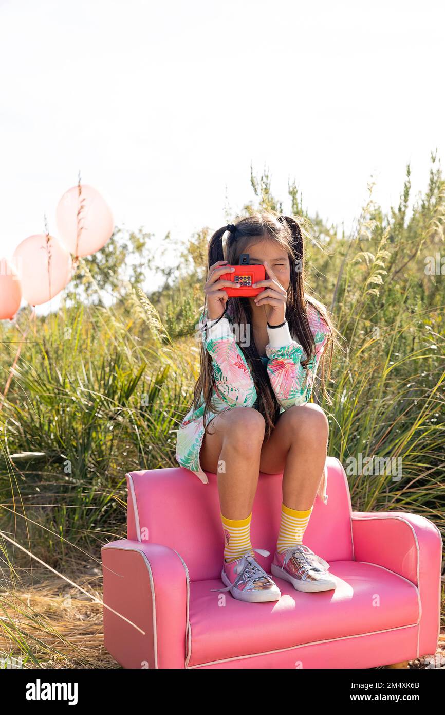 Ragazza fotografando anche se macchina fotografica giocattolo seduta su poltrona rosa Foto Stock