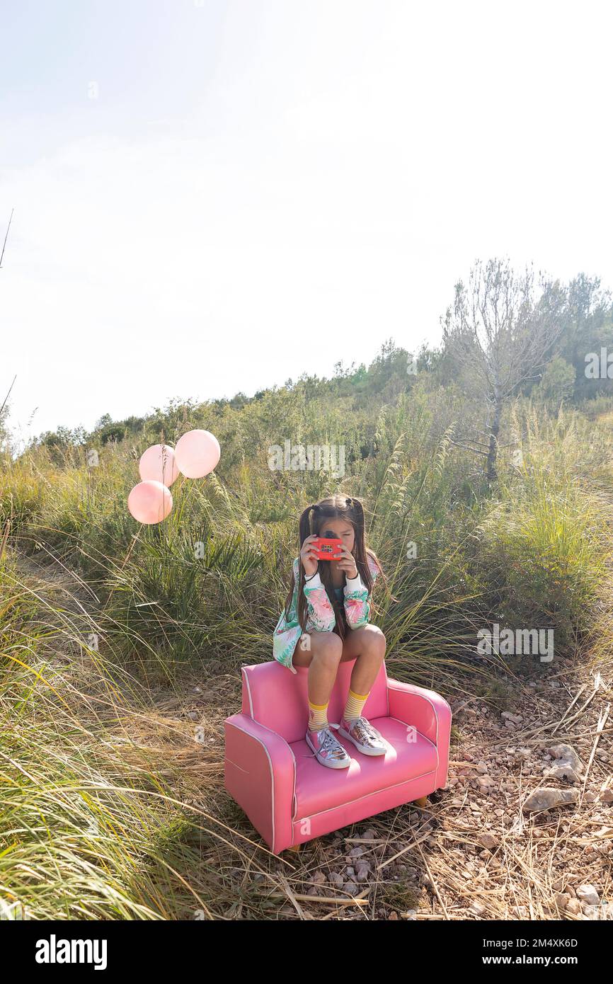 Ragazza che fotografa attraverso una macchina fotografica giocattolo seduta su una poltrona rosa Foto Stock