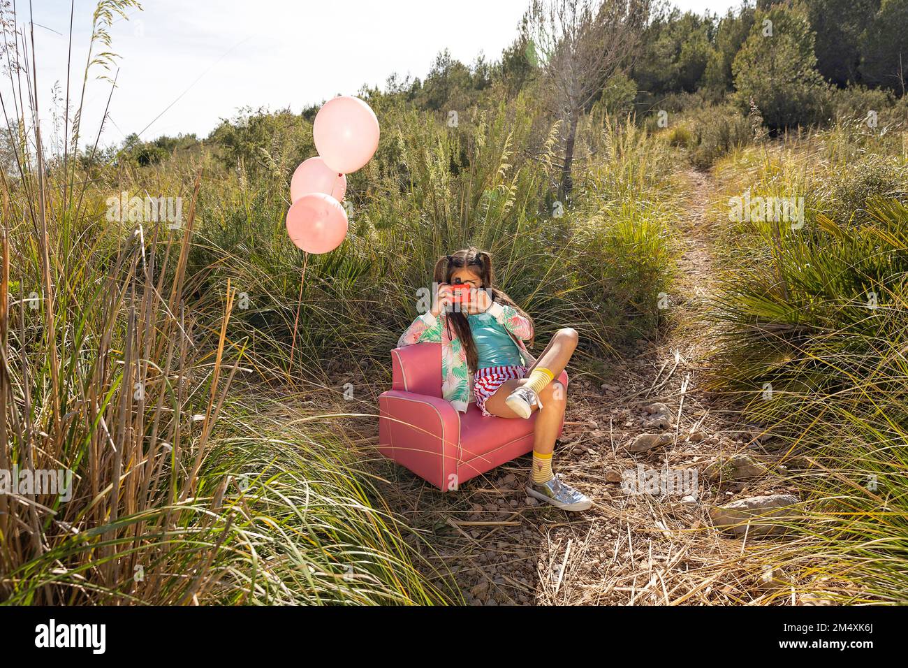 Ragazza che fotografa attraverso macchina fotografica giocattolo seduta su poltrona rosa in campo Foto Stock