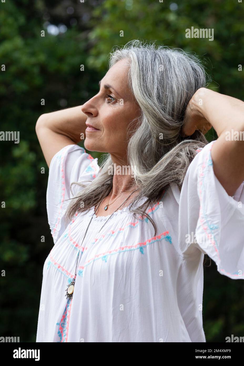 Donna dai capelli grigi che indossa un abito bianco con braccia sollevate nella foresta Foto Stock