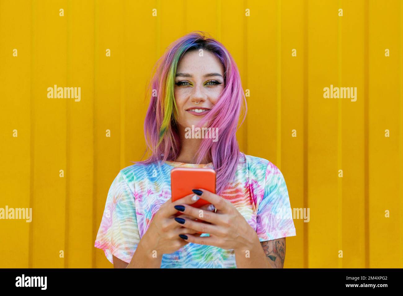 Giovane donna sorridente che tiene il telefono cellulare di fronte al muro giallo Foto Stock