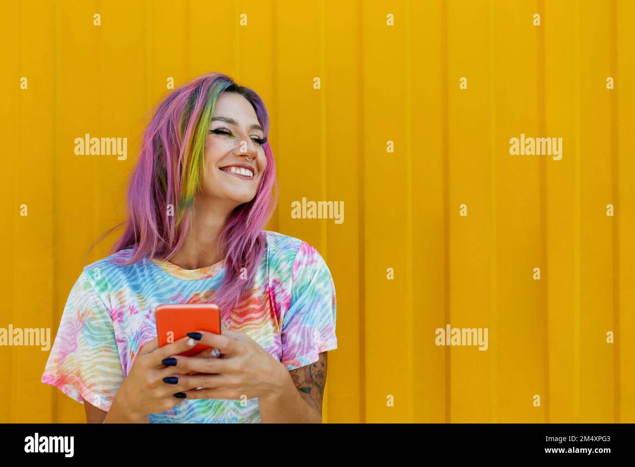 Donna felice con i capelli colorati tenendo il telefono cellulare di fronte alla parete gialla Foto Stock
