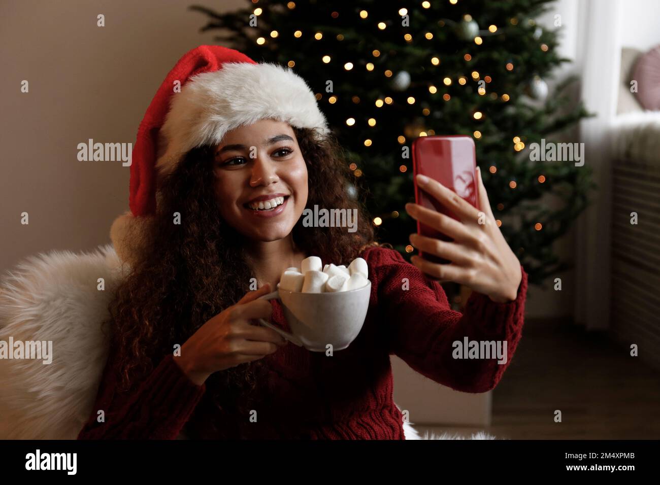 Donna felice che indossa il cappello di Santa e che prende selfie con una tazza di cacao marshmallow Foto Stock