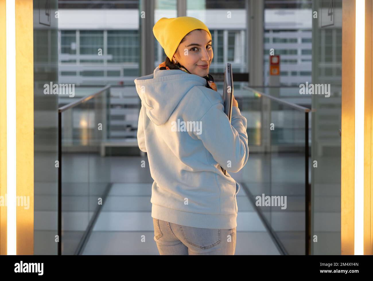 Una studentessa sorridente in una felpa blu e cappuccio giallo cammina lungo il corridoio di vetro Foto Stock