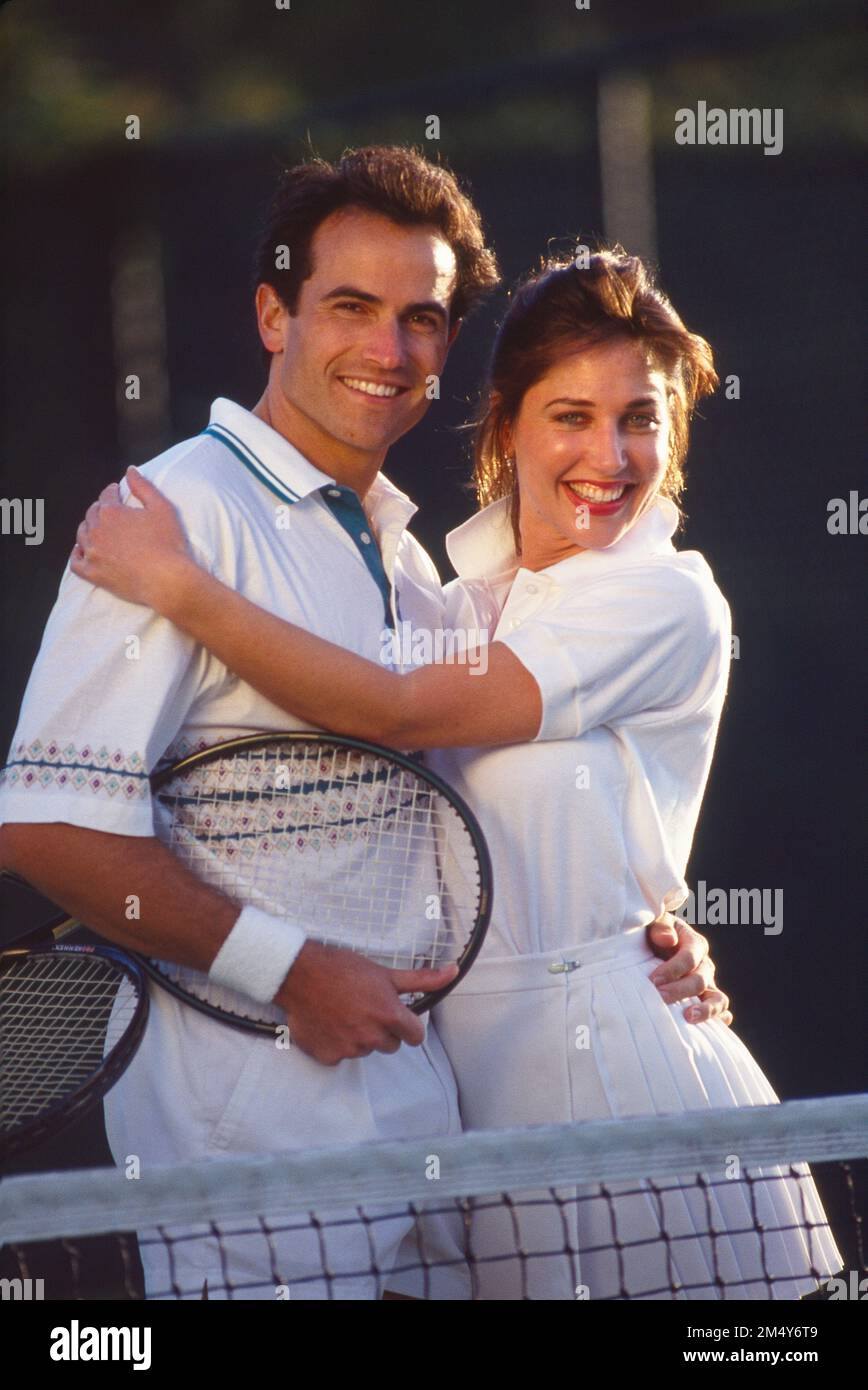 Giovane coppia bianca in abiti da tennis che si posano davanti alla rete, abbracciandosi e sorridendo alla telecamera Foto Stock