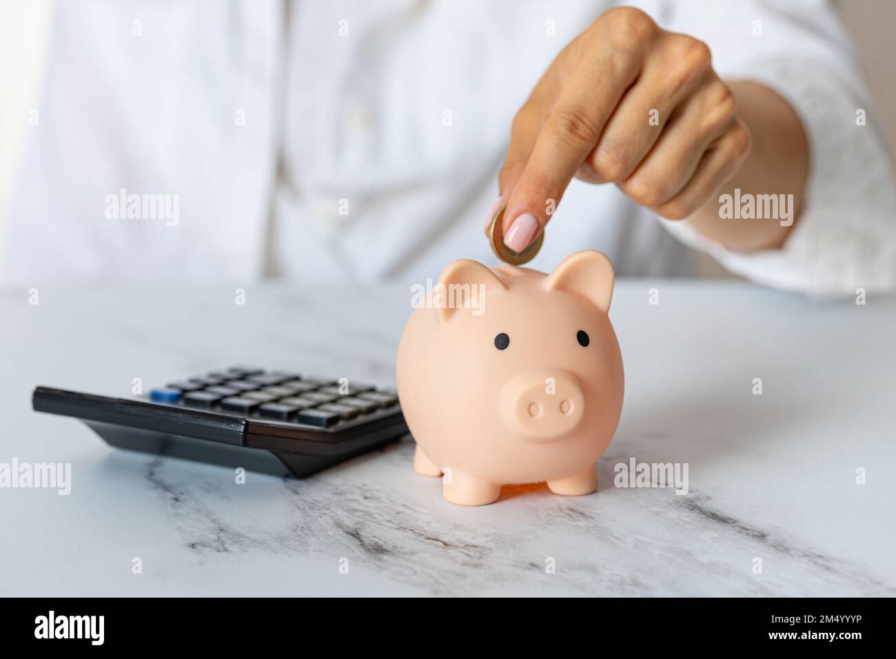 Primo piano delle mani di una donna mettere una moneta in euro in una banca piggy, accanto a una calcolatrice sul tavolo.investimento corretto. Risparmio di denaro in tempi di crisi e. Foto Stock
