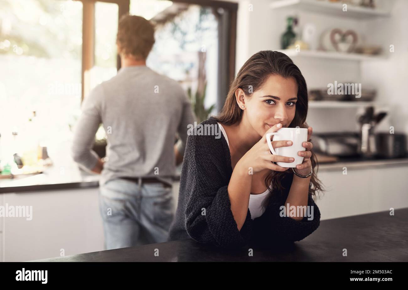 Il caffè mette l'mmm al mattino. Ritratto di una giovane donna che ha il caffè in cucina con il marito in piedi sullo sfondo. Foto Stock