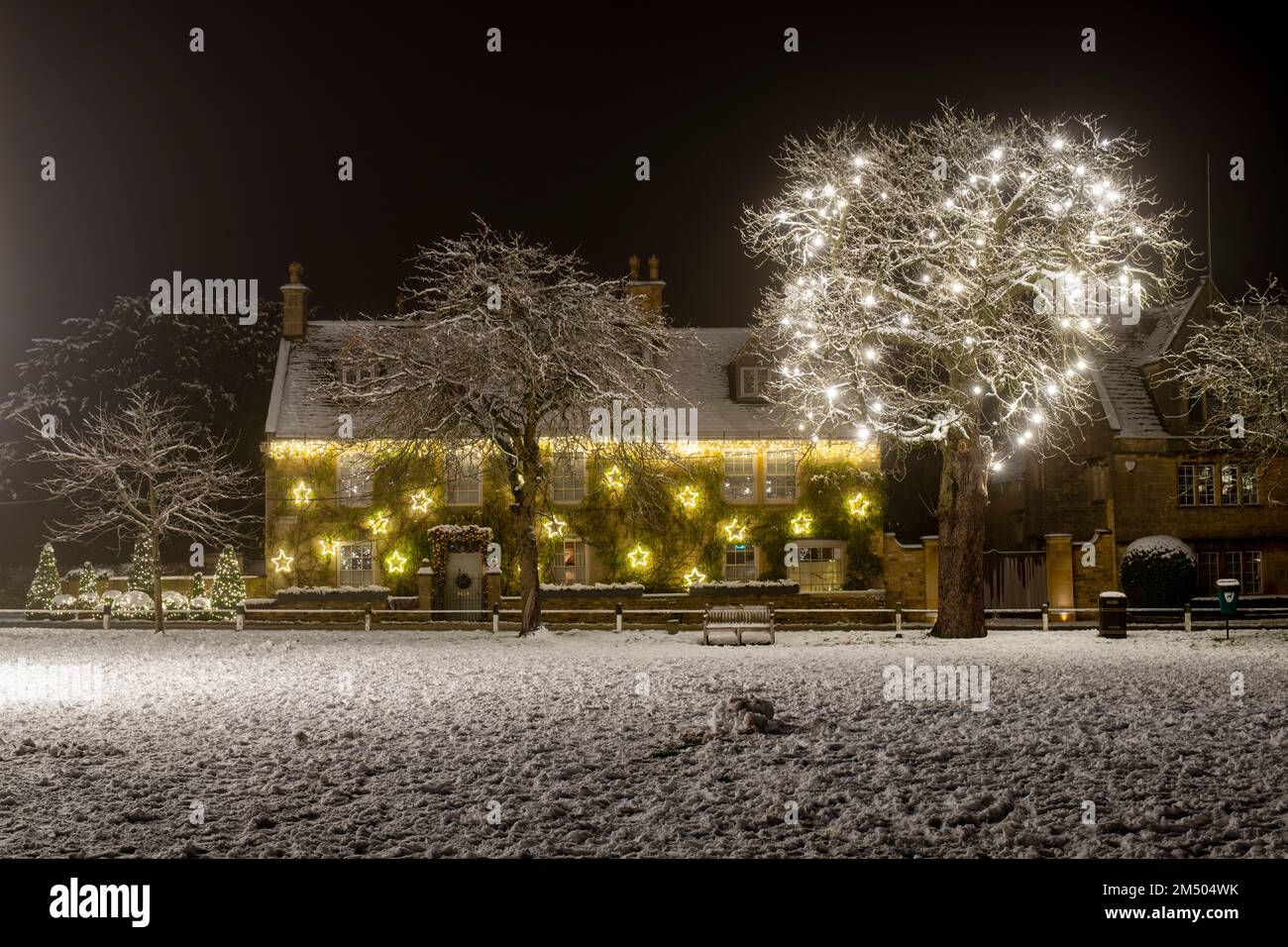 Decorazioni di luce dell'albero di Natale sulla parte anteriore di una casa e negli alberi nella neve di notte. Broadway, Cotswolds, Worcestershire, Inghilterra Foto Stock