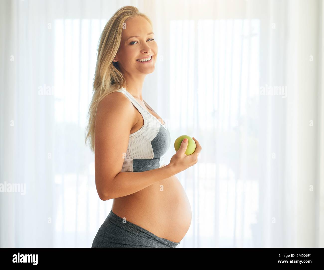 Fare scelte sane per il mio bambino. Ritratto di una donna felice incinta vestita di abbigliamento sportivo e mangiare una mela a casa. Foto Stock