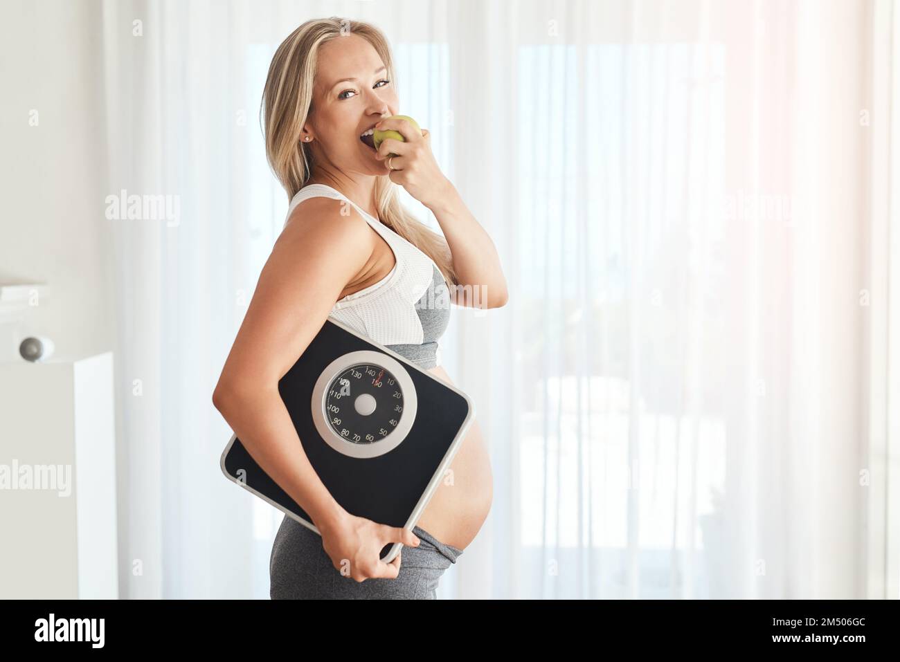 Perché il mio bambino merita un inizio sano nella vita. Ritratto di una donna felice incinta vestita di abbigliamento sportivo e che porta una bilancia mentre mangia una mela Foto Stock