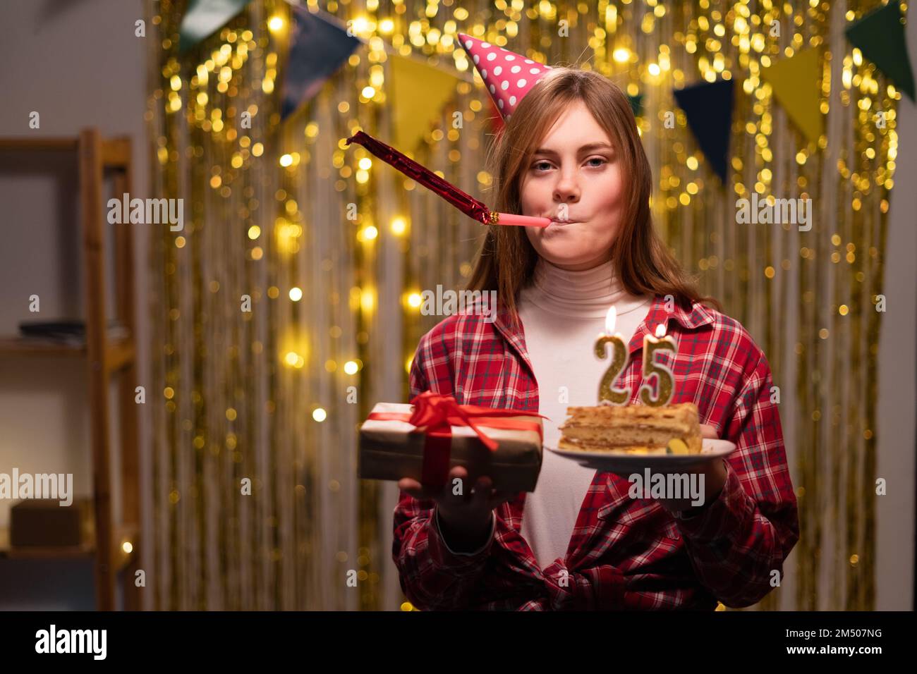 Festa di compleanno. Giovane donna con regalo e torta di compleanno con candele numero 25. Festa di compleanno a casa. Cappello da festa in testa. Spazio di copia Foto Stock