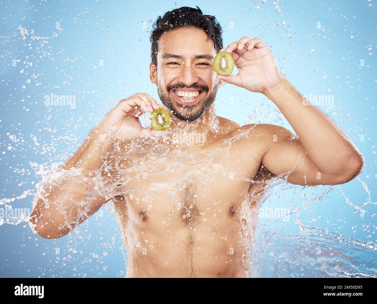 Ritratto facciale, spruzzi d'acqua e uomo con kiwi in studio isolato su sfondo blu. Pulizia, cura della pelle e igiene di felice modello maschile lavaggio Foto Stock