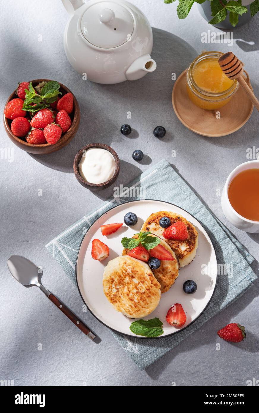 Frittelle con formaggio casolare o sciroppi con blerries freschi, miele e panna acida su sfondo blu con una tazza di tè e un bollitore. Il concetto di Foto Stock