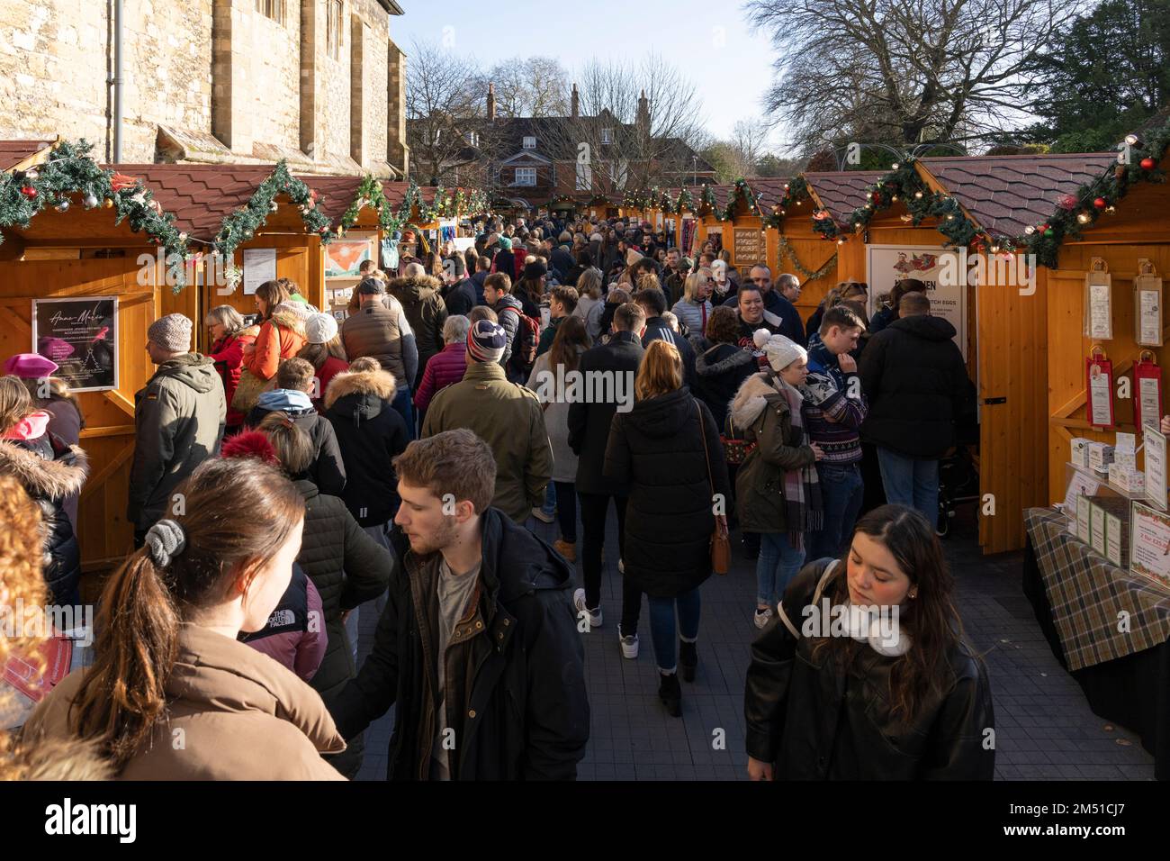 Gli amanti dello shopping natalizio al mercatino di Natale di Winchester con chalet in legno accanto alle mura medievali storiche della cattedrale gotica di Winchester, Inghilterra, Regno Unito Foto Stock