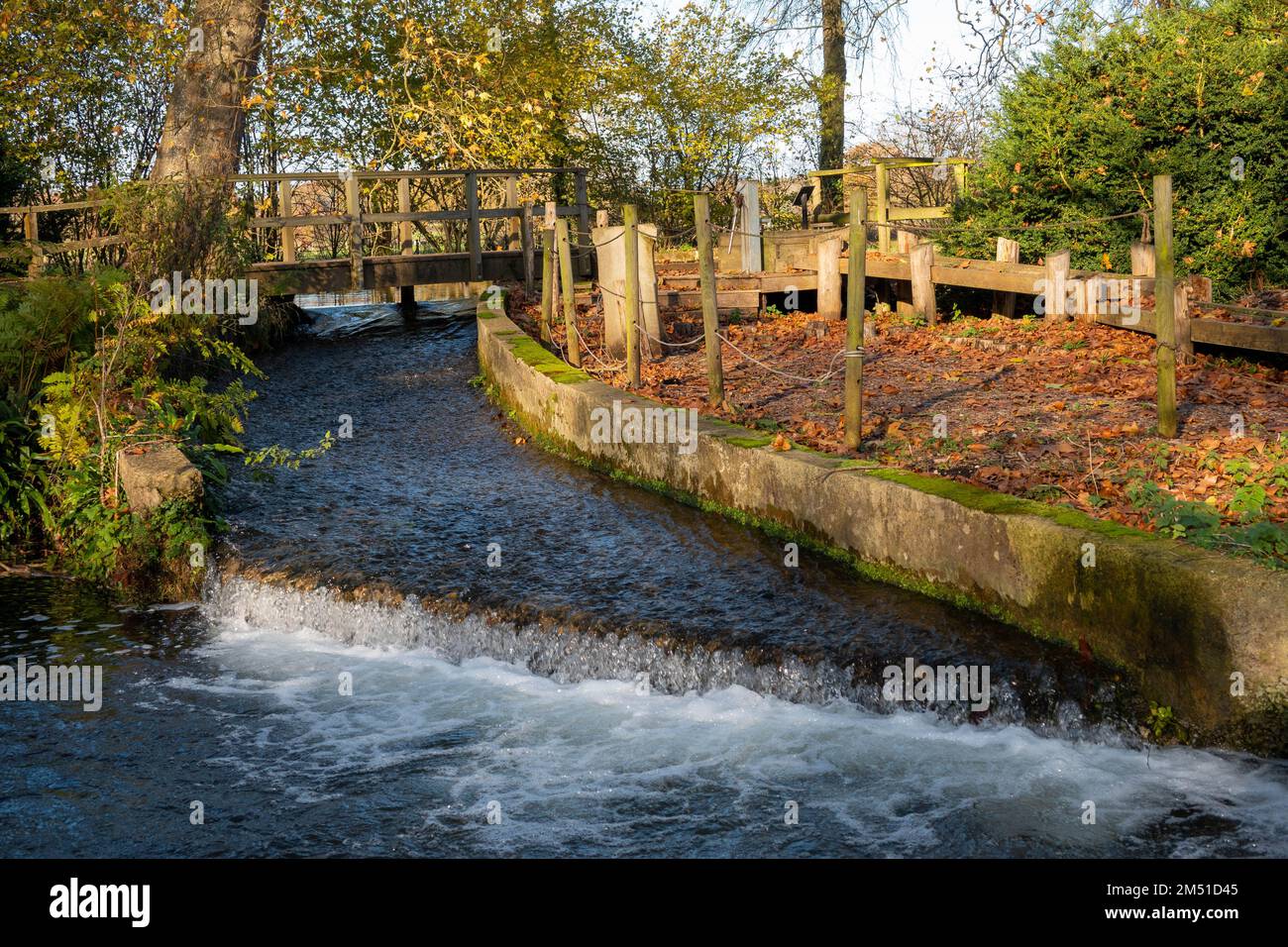 acqua che cade su un piccolo stramazzo nel fiume con un vecchio ponte di legno sullo sfondo Foto Stock