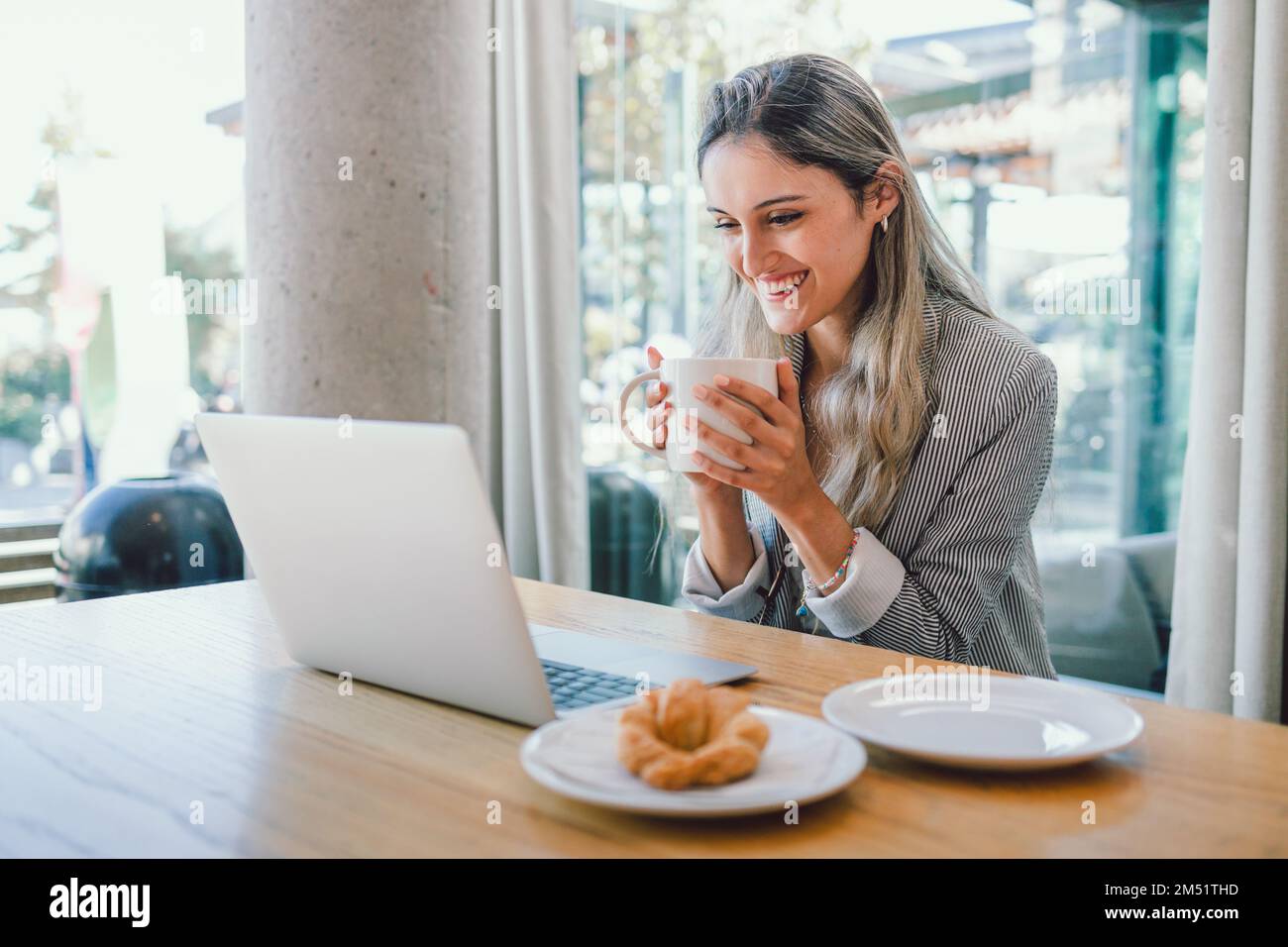 Giovane donna d'affari felice che usa un computer portatile, bevendo tè o caffè da una tazza in un moderno ambiente di lavoro aperto Foto Stock
