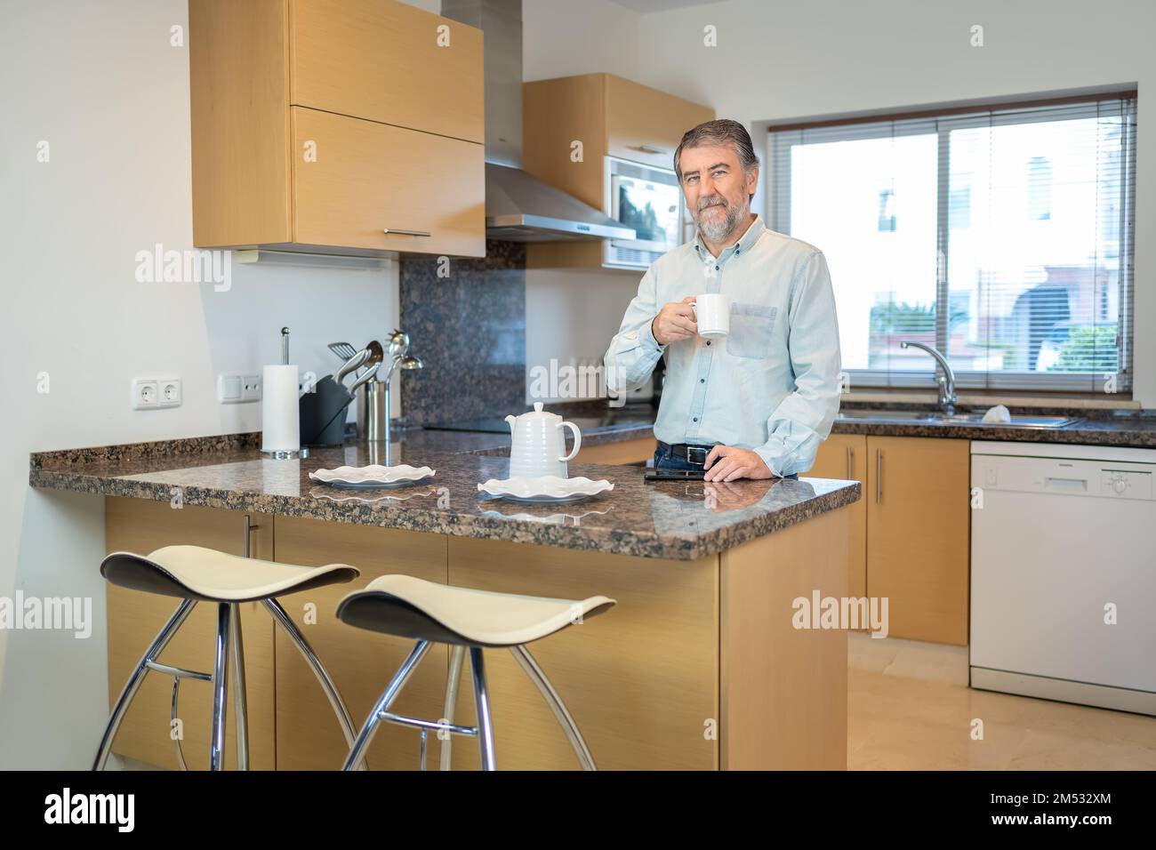 Uomo bianco anziano che ha una tazza di caffè in cucina a casa e guardando la macchina fotografica. Foto Stock