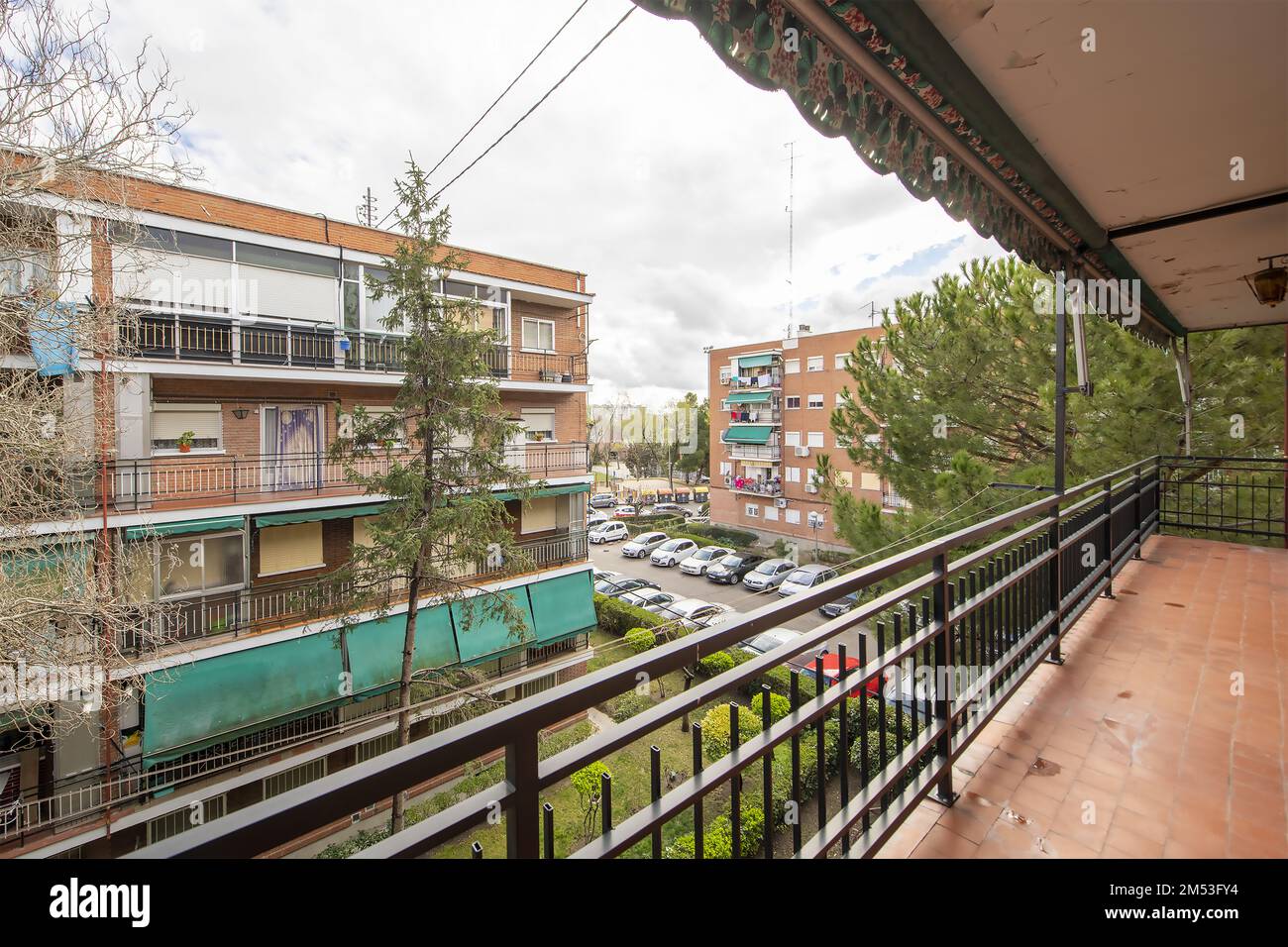 Lunga terrazza di una casa con una ringhiera di metallo nero, tende raccolte sul tetto e vista di aree comuni con giardini e siepi Foto Stock