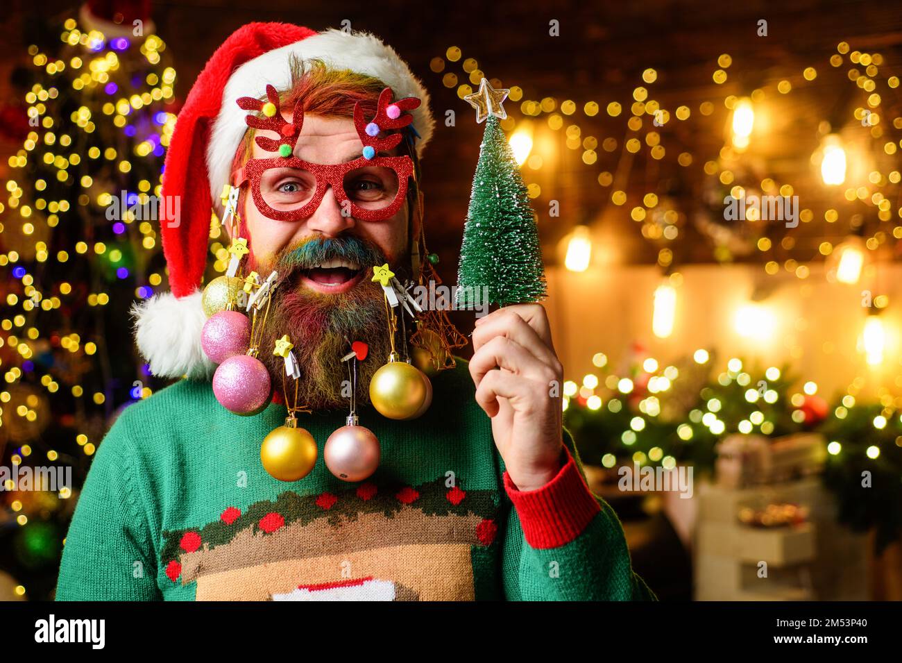 Barbuto uomo in cappello di Santa e bicchieri da festa con piccolo albero di Natale. Barba decorata per Capodanno. Foto Stock