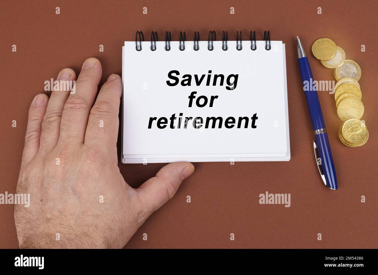 Concetto aziendale. Su una superficie marrone ci sono monete, una penna e un blocco note con l'iscrizione - risparmio per pensionamento. Il notebook sta tenendo una mano. Foto Stock