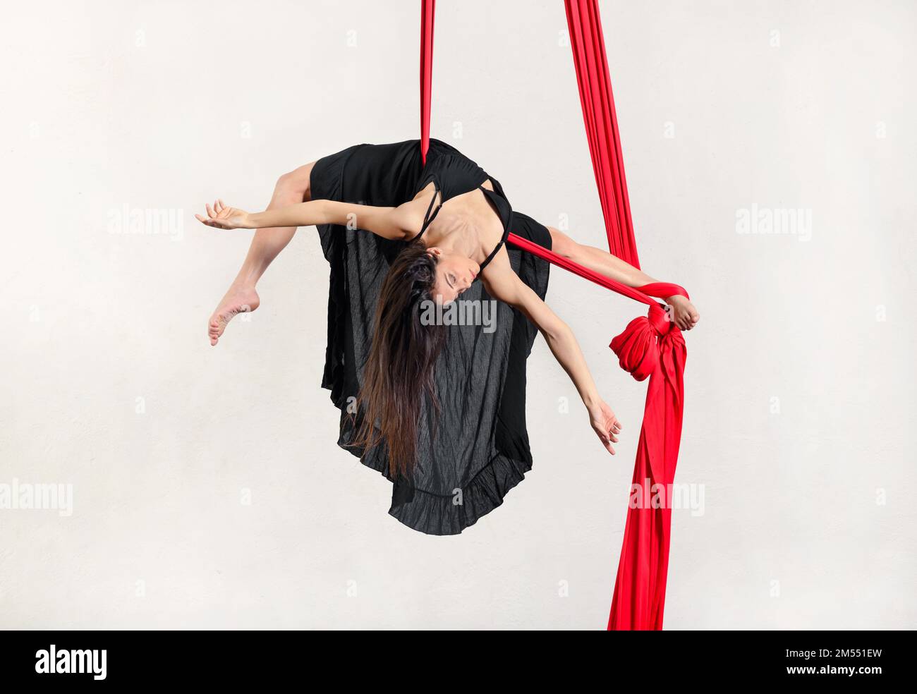 Corpo pieno di ballerina giovane e graziosa barefooted che si esibisce ad arco su sete aeree rosse appese su sfondo bianco Foto Stock