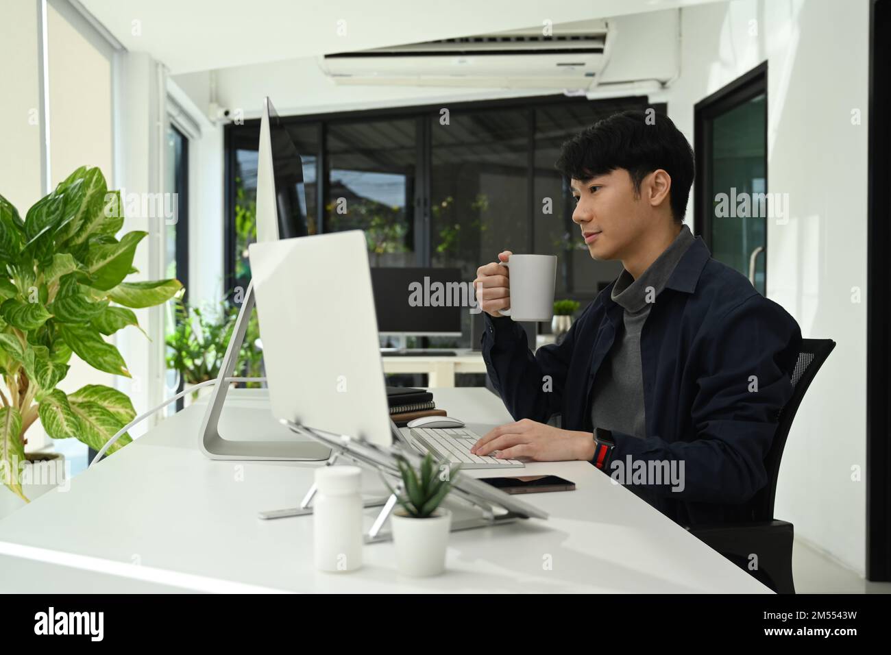 Uomo d'affari asiatico sorridente bere caffè e leggere e-mail di lavoro sullo schermo del computer Foto Stock