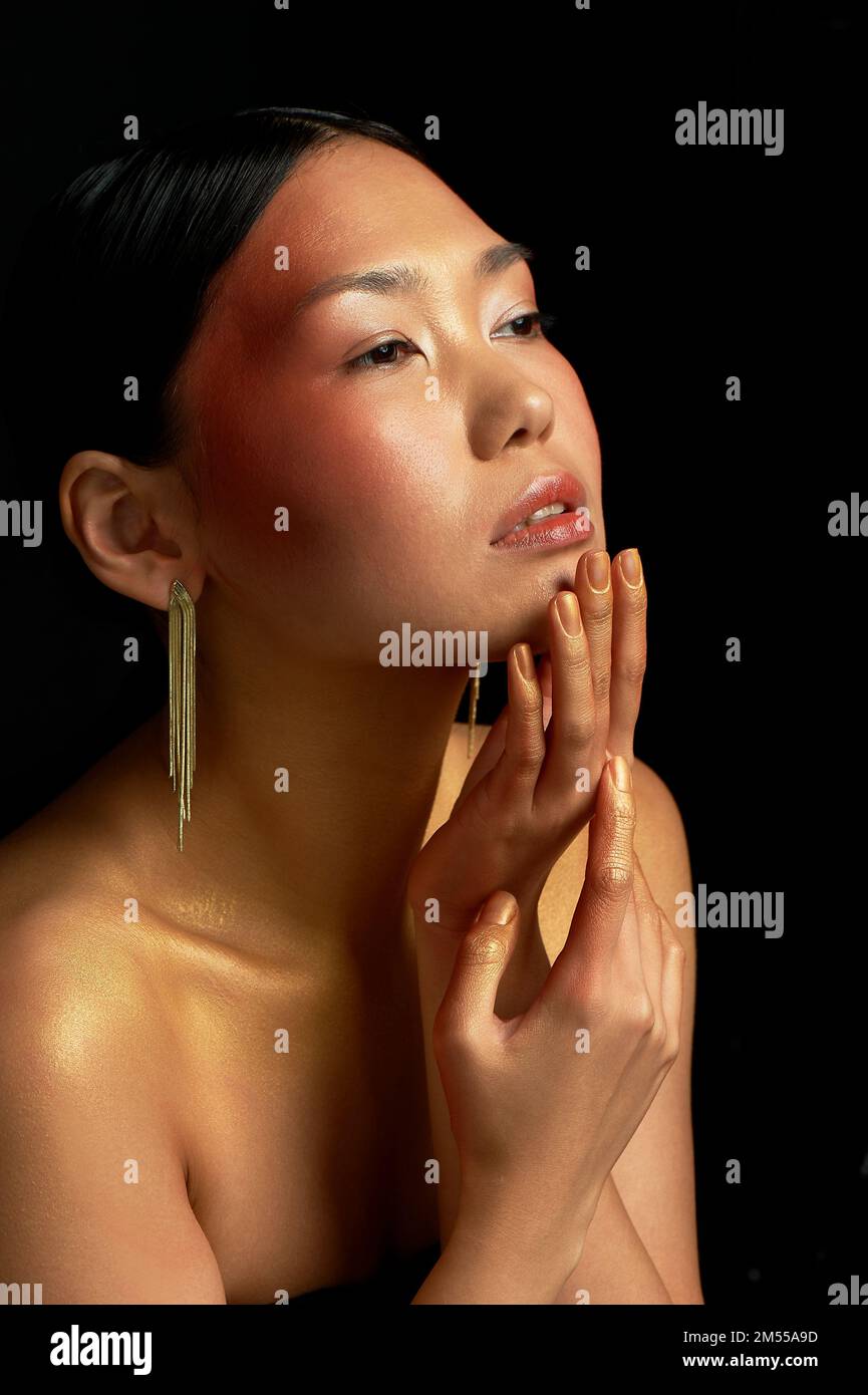 Una ragazza asiatica su sfondo nero in studio. Oro. Collarbones sottili. Il regno della bellezza e del profumo. Tocca delicatamente il viso Foto Stock