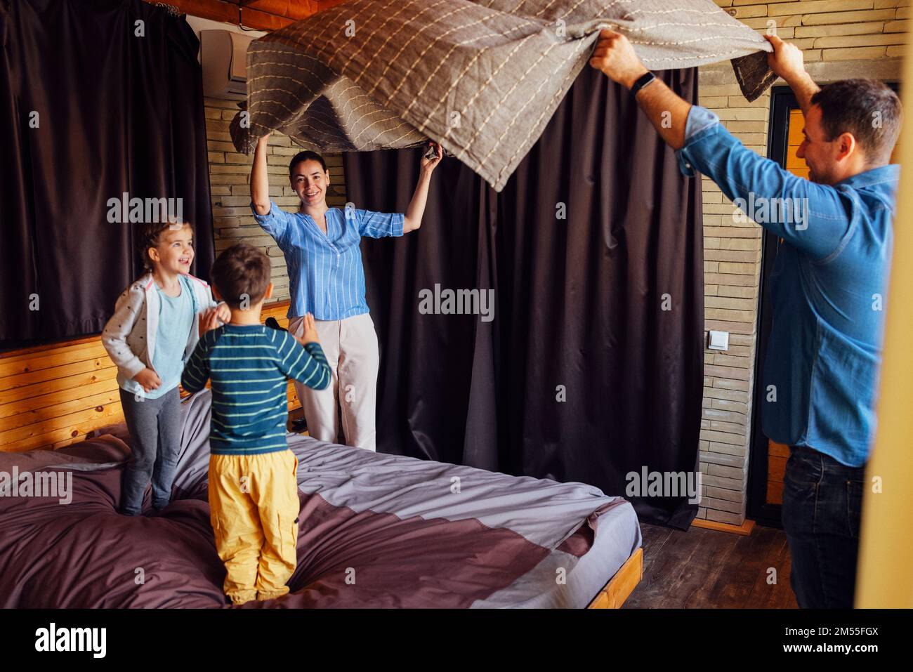 Famiglia felice madre multietnica, padre e bambini, figlio e figlia, ridendo, giocando, combatte i cuscini e salta a letto in camera da letto a casa in corride Foto Stock