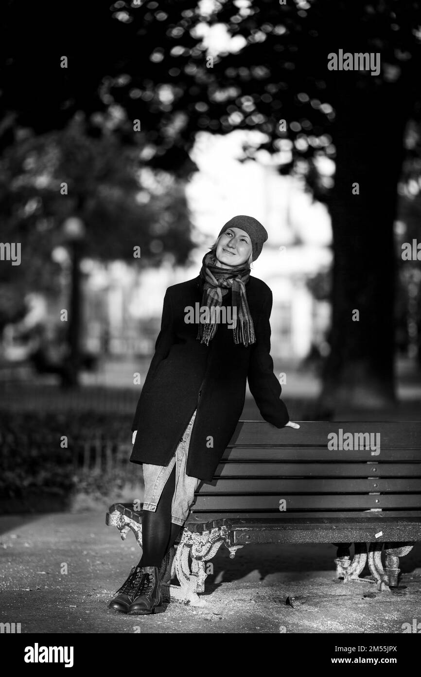 Una donna in un cappotto autunnale in posa vicino ad una panca del parco. Foto in bianco e nero. Foto Stock