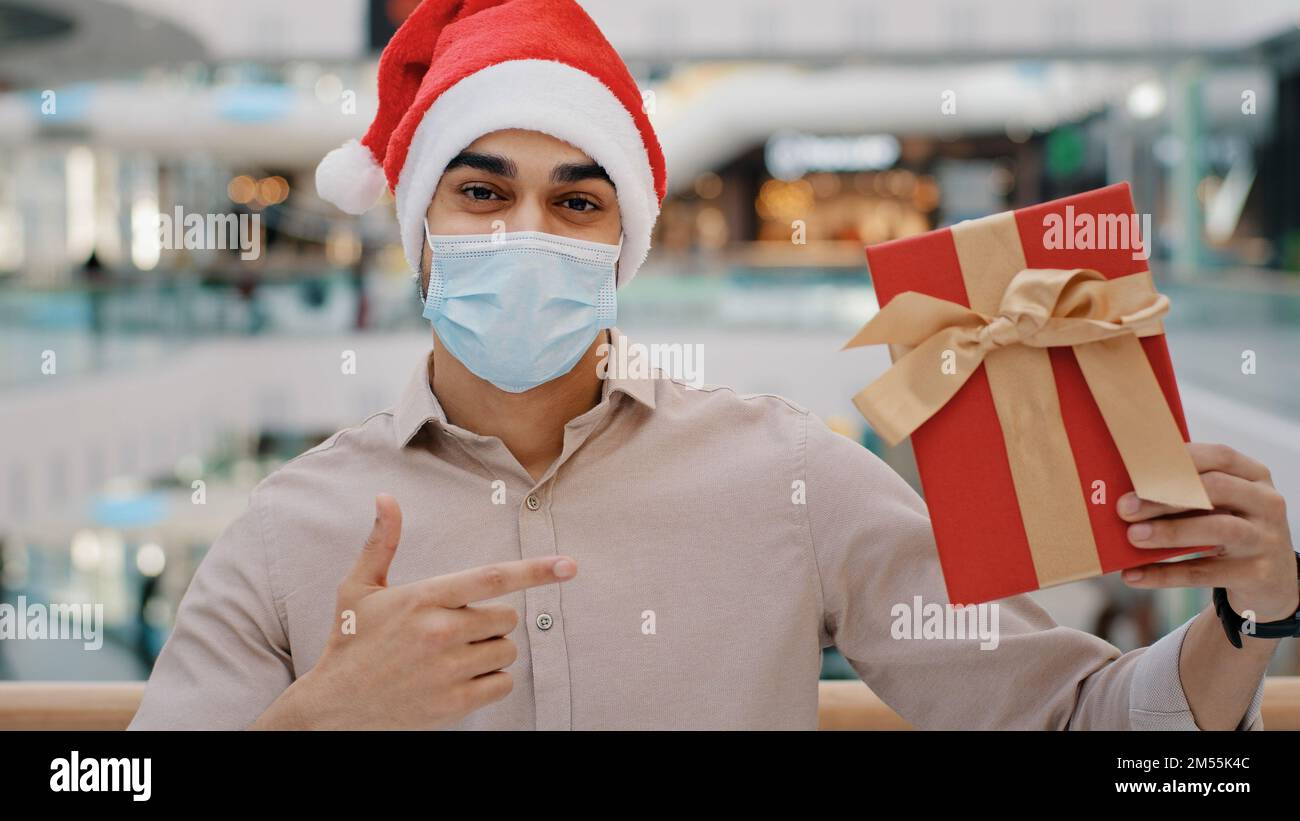Uomo in Natale X-mas cappello ragazzo maschio in maschera di fronte Covid in piedi nel centro commerciale centro commerciale guardando la macchina fotografica che mostra regalo scatola di regalo avvolto in rosso Foto Stock
