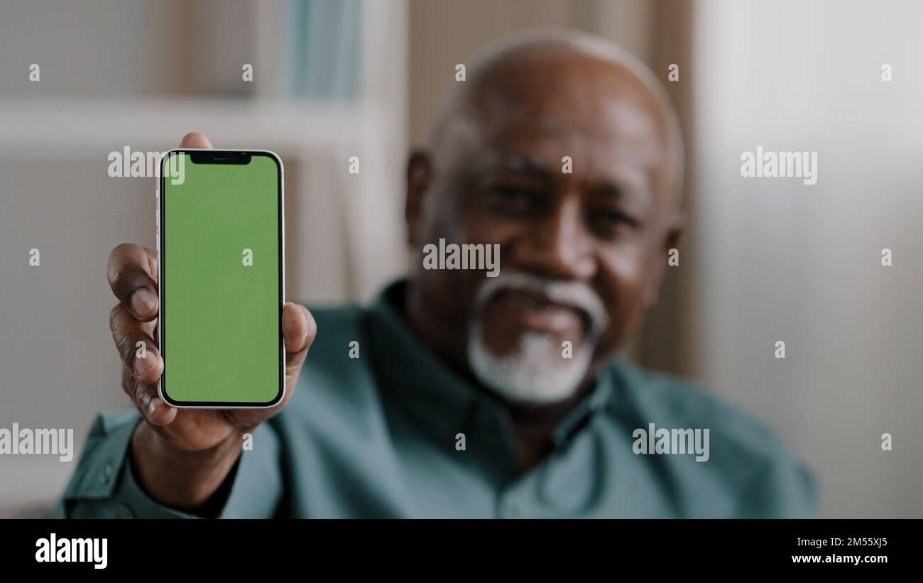 Vecchio uomo afro-americano biracial a casa seduto sul divano uomo d'affari in ufficio mostrando smartphone con schermo verde vuoto chromakey tenere Foto Stock