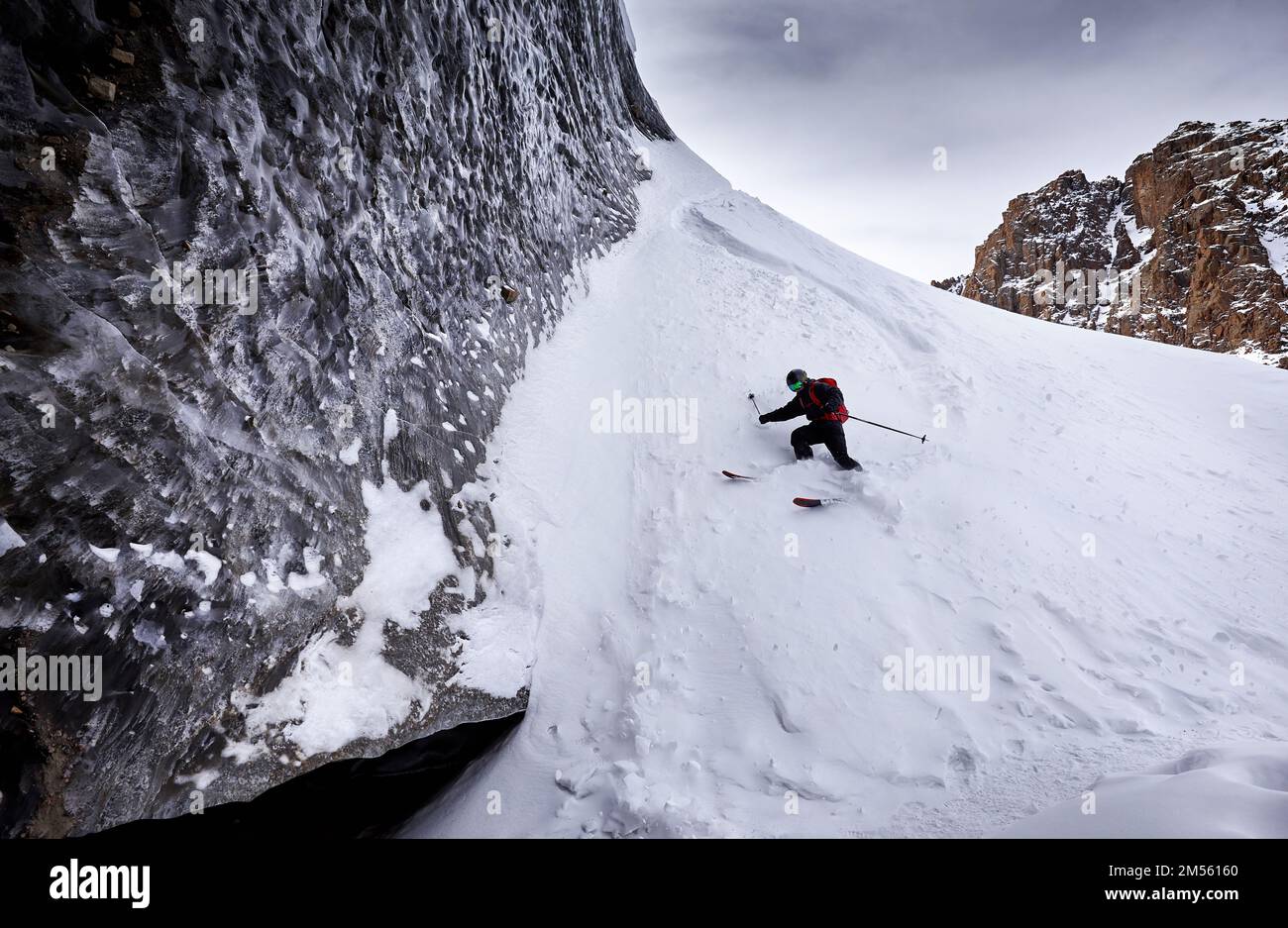 Sciatore uomo sciare in discesa con zaino rosso in casco e maschera in alte montagne innevate vicino al muro di ghiaccio con spruzzi di neve. Sport estremi all'aperto in wint Foto Stock