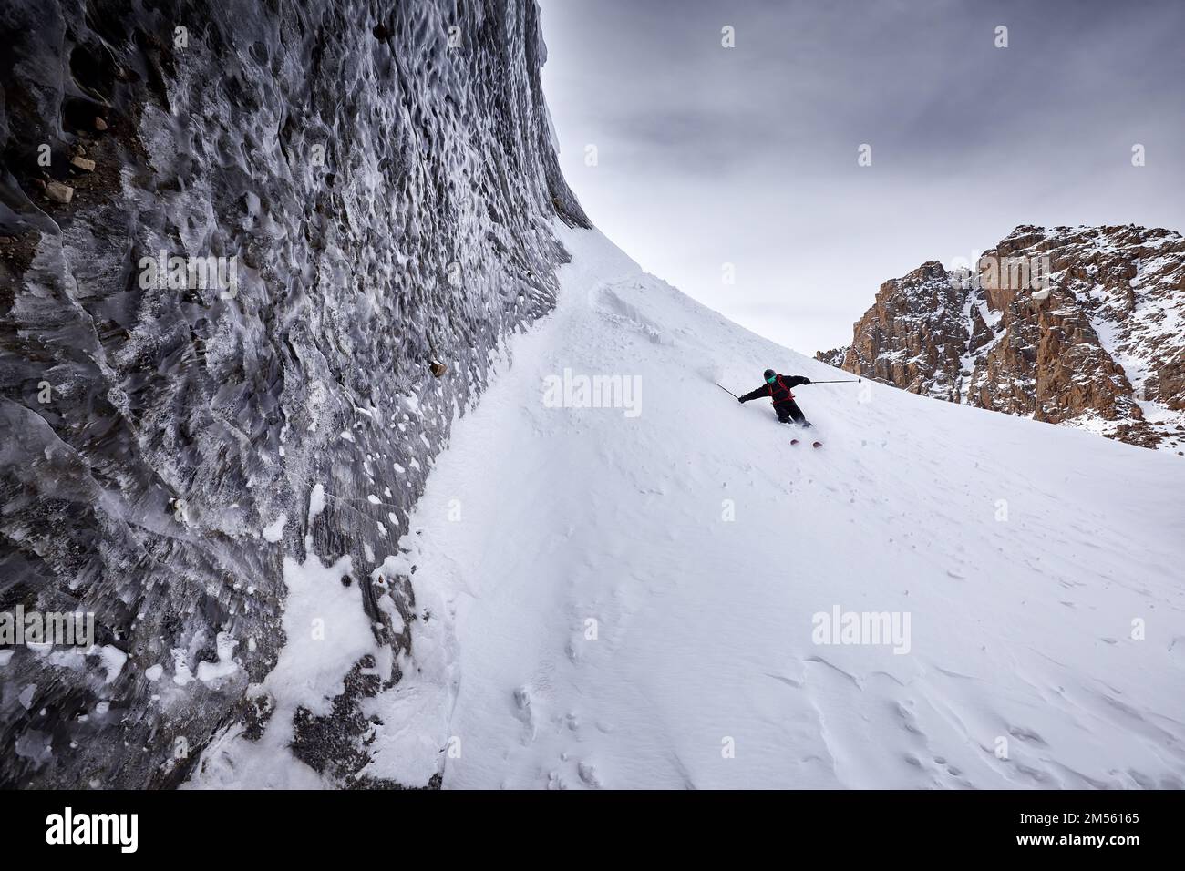 Sciatore uomo sciare in discesa con zaino rosso in casco e maschera in alte montagne innevate vicino al muro di ghiaccio con spruzzi di neve. Sport estremi all'aperto in wint Foto Stock