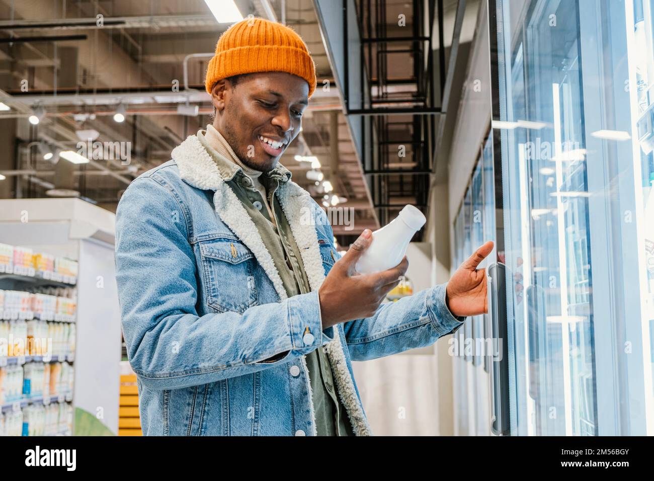 Il ragazzo afro-americano positivo in giacca di denim e cappello arancione prende la bottiglia di latte dalla scatola del frigorifero in vista laterale del moderno supermercato Foto Stock